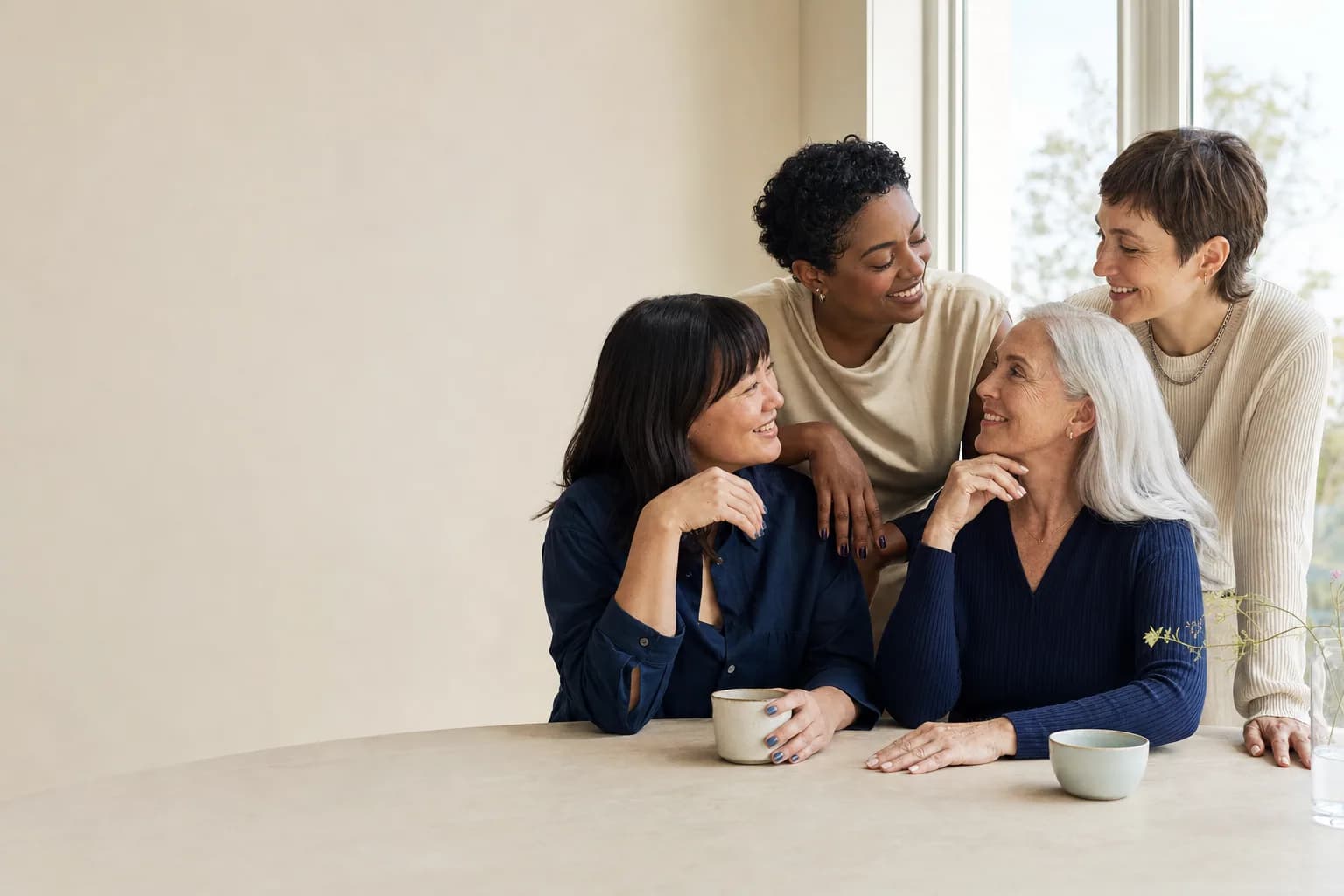 Four adults of different ages and skin tones gather closely around a pale table by a bright window, smiling together over cups of tea, several with softly painted nails.